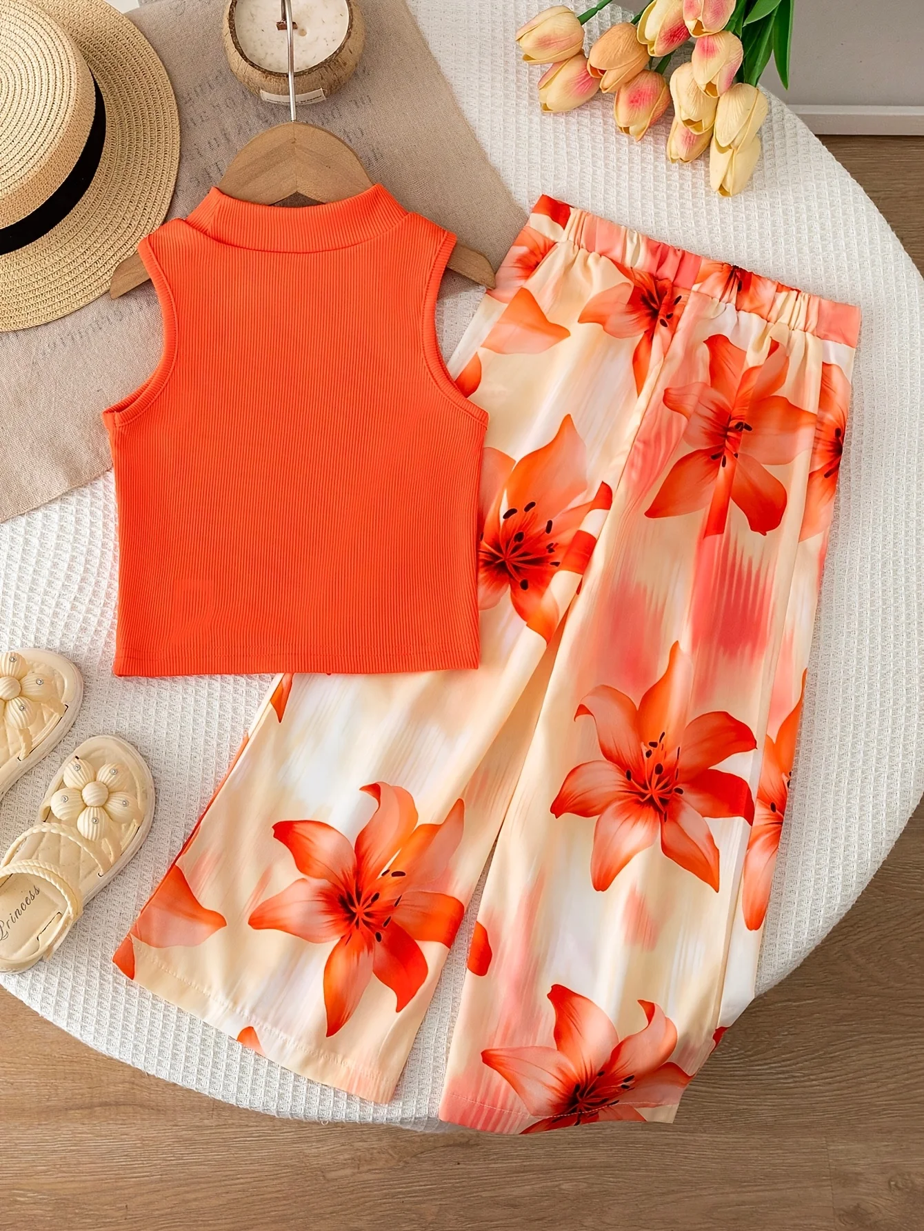 Conjunto de dos piezas estilo vacacional naranja para niñas: top corto sin mangas con cuello simulado, de color liso y textura acanalada, combinado con pantalón fluido con estampado de lirios.